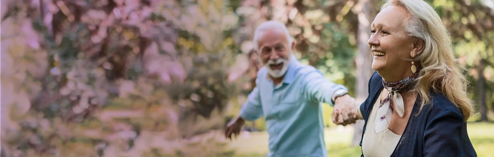 elderly couple hold hands and walk through an orchard, smiling